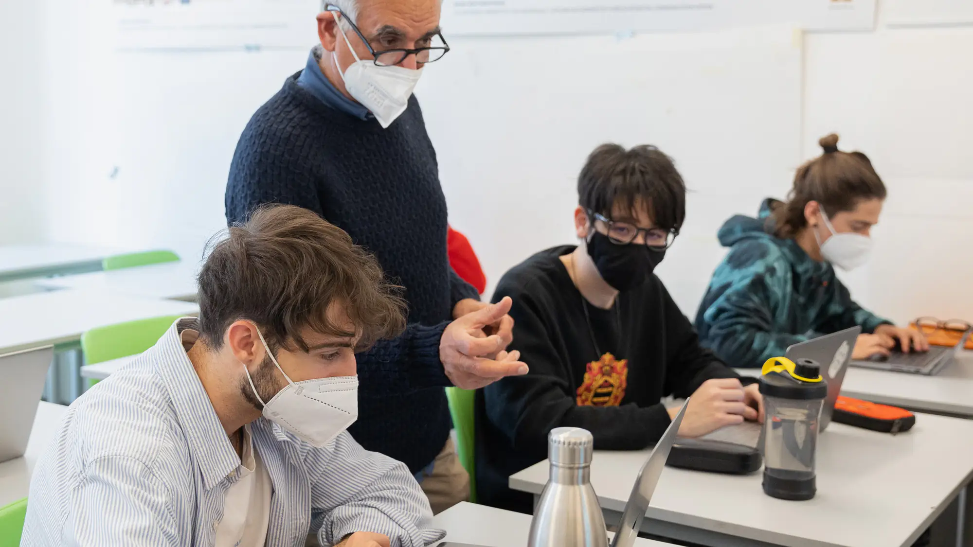 A Professor wearing a face mask supervises students working on laptops at IAAD University during a Master in Digital Marketing class